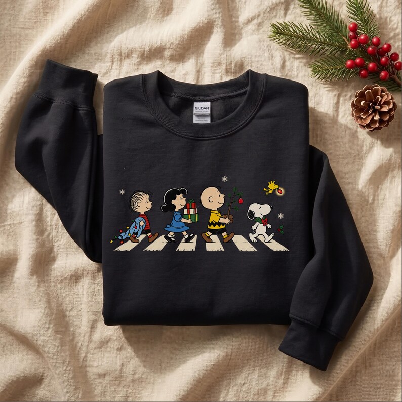 May include: A black long-sleeve sweatshirt featuring a graphic print of the Peanuts characters Linus, Lucy, Charlie Brown, Snoopy, and Woodstock walking across a crosswalk, carrying Christmas gifts and a small tree with snowflakes.