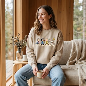 May include: A beige crewneck sweatshirt featuring a graphic print of the Peanuts characters walking in a line. The design shows Linus, Lucy, Charlie Brown, and Snoopy carrying various items, with Woodstock flying above them on a white background.
