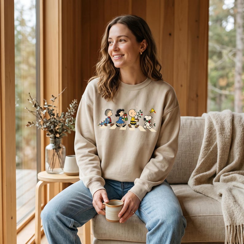 May include: A beige crewneck sweatshirt featuring a graphic print of the Peanuts characters walking in a line. The design shows Linus, Lucy, Charlie Brown, and Snoopy carrying various items, with Woodstock flying above them on a white background.