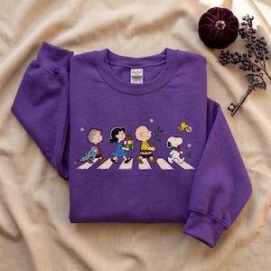May include: A purple long-sleeve sweatshirt featuring a graphic print of Peanuts characters walking across a crosswalk. The design includes Linus, Lucy, Charlie Brown, Snoopy, and Woodstock with holiday items like gifts and a small decorated tree.