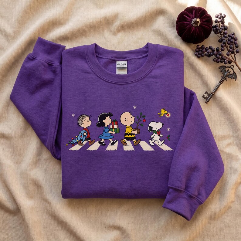 May include: A purple long-sleeve sweatshirt featuring a graphic print of Peanuts characters walking across a crosswalk. The design includes Linus, Lucy, Charlie Brown, Snoopy, and Woodstock with holiday items like gifts and a small decorated tree.
