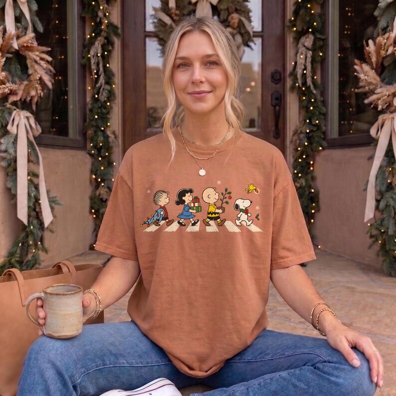 May include: A tan short-sleeve t-shirt featuring a graphic of four Peanuts characters walking in a line across a white crosswalk. The characters include Linus, Lucy, Charlie Brown, and Snoopy, each carrying holiday items like gifts and a tree.