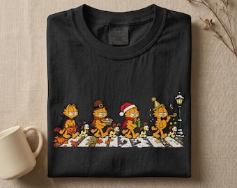 May include: A black t-shirt featuring a graphic of Garfield and Odie walking across a crosswalk in seasonal outfits. The characters transition from Halloween costumes to Thanksgiving, Christmas, and New Year's attire. The Comfort Colors label is visible.