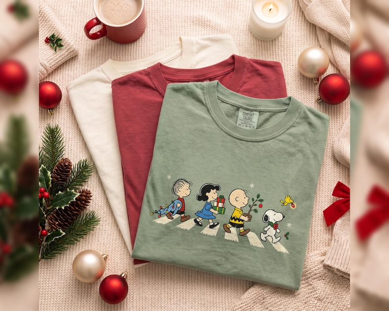 May include: Three crew neck t-shirts in cream, maroon, and sage green. The sage green shirt features a graphic print of the Peanuts characters walking in a line, carrying Christmas items, with the Comfort Colors brand label visible at the neck.