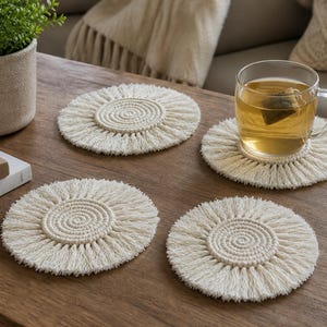 Könnte beinhalten: Ein Set aus vier runden, cremeweißen Makramee-Untersetzern mit einem zentralen Spiralmuster aus geknoteter Kordel, umgeben von einem Fransenrand. Ein Untersetzer trägt eine klare Glastasse, die mit Tee und einem Teebeutel gefüllt ist.
