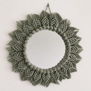 May include: A circular wall mirror framed by sage green macrame cord. The frame features a repeating pattern of knotted leaf-like shapes and fringe, creating a sunburst effect. A single cord loop at the top allows for hanging the mirror on a wall.