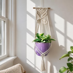 Può includere: Un portapiante in macramè color crema sospeso a un bastone di legno orizzontale, caratterizzato da intricati motivi a nodi. Il supporto sostiene un vaso in ceramica viola lucido con una pianta verde, terminato da una lunga nappa.