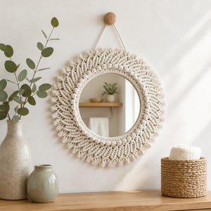 Op de afbeelding: Een ronde wandspiegel met een ingewikkeld gebroken wit macramé frame met geknoopte patronen en een gefranjerde rand. De spiegel hangt aan een koord aan een houten knop en toont een reflecterend glazen oppervlak met een interieur op de achtergrond.