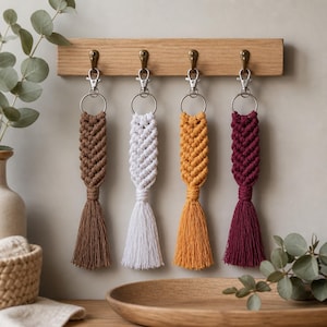 Pode incluir: Quatro porta-chaves de macramé pendurados num suporte de madeira. Cada porta-chaves possui um mosquetão de metal prateado, uma argola, uma secção de cordão entrançado em castanho, branco, amarelo mostarda ou bordeaux e uma borla.