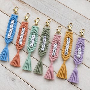 Peut inclure: Sept porte-clés en macramé bleu, orange, vert clair, vert olive, rose, jaune et violet. Chaque porte-clés comporte un mousqueton doré, une rangée centrale verticale de perles carrées blanches avec des lettres noires formant des prénoms.