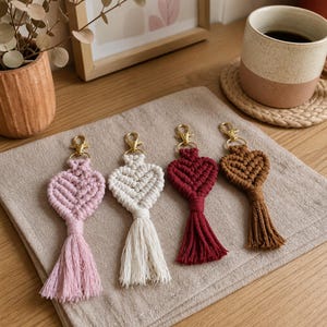 Peut inclure: Quatre porte-clés en macramé en forme de cœur avec des fermoirs mousquetons dorés. Les porte-clés sont alignés et présentent des couleurs rose clair, blanc cassé, rouge foncé et marron. Chaque cœur est doté d'un pompon assorti en bas.