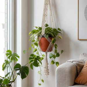 Op de afbeelding: Een crèmekleurige macramé plantenhanger met ingewikkelde knooppatronen en een lange kwast aan de onderkant. De hanger bevat een terracotta-kleurige pot met een pothosplant met bonte, hartvormige bladeren in groen en geel.