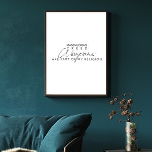 May include: A framed print with a white background and black text that reads "MANDALORIAN CREED Weapons ARE PART OF MY RELIGION". The print is hanging on a teal wall above a teal couch with a brown throw blanket.