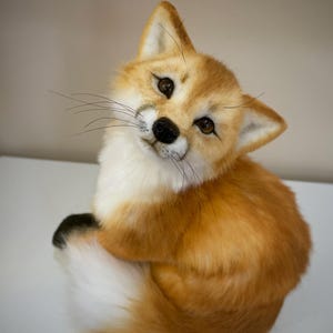 May include: A handcrafted plush fox figurine with realistic orange and white fur, dark brown eyes, a black nose, and long thin whiskers. The fox is in a seated position with its bushy tail curled around its body, featuring a black tip on the tail.