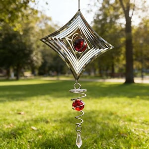 Könnte beinhalten: Ein Windspiel aus silberfarbenem Metall mit einem rautenförmigen Rahmen und konzentrischen quadratischen Ausschnitten. Eine rote Glaskugel hängt in der Mitte. Darunter befindet sich eine Metallspirale mit einer zweiten roten Kugel und einem Kristall.