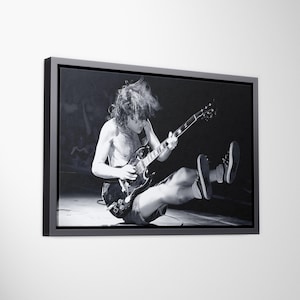 May include: A black and white framed canvas print depicting a musician with long hair playing an electric guitar while jumping in the air. The performer is shirtless and wearing shorts and sneakers, captured in a dynamic stage performance pose.