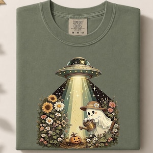 May include: An olive green t-shirt featuring a graphic of a ghost wearing a straw hat and watering a small pumpkin. Above the ghost, a UFO emits a bright beam of light over a garden of sunflowers and daisies. The shirt has a Comfort Colors label.