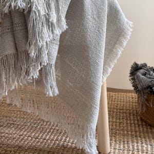 May include: A light gray woven throw blanket featuring a textured, open-weave fabric construction with long, loose fringe detailing along the edges. The textile displays a natural, slightly uneven appearance with visible horizontal and vertical threads.