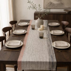 May include: A dark wood dining table set for six with white plates, folded beige napkins, and gold-toned cutlery. A long table runner with orange and blue-grey stripes runs down the center, featuring a ceramic vase with branches and two candles.