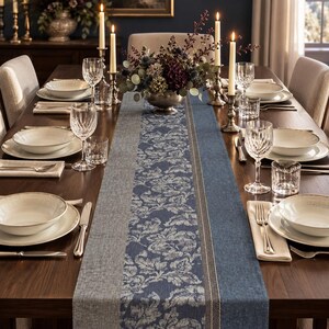 Puede incluir: Una mesa de comedor larga preparada con un camino de mesa floral en azul y gris. El servicio incluye cuencos de cerámica blanca sobre platos, cristalería transparente, cubiertos de plata, servilletas plegadas y velas altas en candelabros.