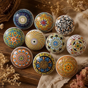 Peut inclure: Une collection de neuf boutons de porte ronds en céramique présentant des motifs géométriques et floraux variés, complexes et colorés dans des tons de bleu, jaune, orange, rouge, noir et blanc, disposés sur une surface en bois avec du tissu.