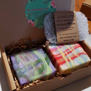 May include: A gift box containing two bars of handmade soap and a soap lift. One soap bar is purple and green, the other is red, yellow, and blue. The soap lift is made of eco-friendly bio-plastic. The box has a floral logo.