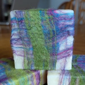 May include: Three bars of soap wrapped in colorful, hand-felted wool. The wool is a mix of green, blue, purple, and white.