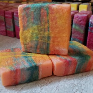 May include: Three square bars of handmade soap with a vibrant, colorful design. The soap features layers of orange, yellow, blue, and red hues. Other bars of soap in various colors are visible in the background.
