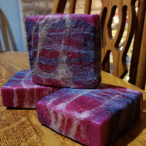 May include: Three handmade soap bars with a vibrant pink base, overlaid with blue and white fibers. The soaps are rectangular and stacked on a wooden surface, showcasing a textured, artistic design. The soaps appear to be felted.