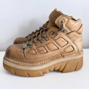 Pode incluir: Uma bota de cano curto em couro bege com uma sola plataforma grossa de borracha com sulcos profundos. A bota apresenta uma silhueta de cano alto, ilhós metálicos em forma de D para os atacadores e um colarinho acolchoado.
