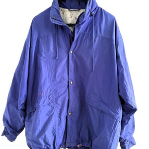 May include: A purple windbreaker jacket featuring a high collar with a drawstring, a front zipper closure with snap buttons, and long sleeves. The interior lining displays a white fabric with a red graphic print pattern throughout the garment.