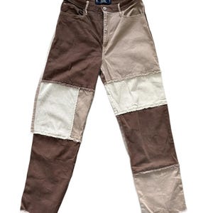 May include: High-waisted trousers featuring a patchwork design in shades of dark brown, light brown, and cream. The garment includes a button and zip fly closure, front pockets, and visible frayed seams along the horizontal panel edges.