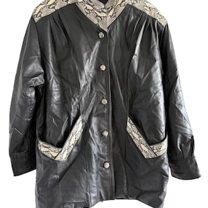 May include: A black leather jacket featuring a stand-up collar and shoulder panels with a beige and black snakeskin print. The jacket has a front button closure with five silver-toned buttons and two front pockets trimmed with snakeskin print.
