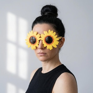 May include: Novelty sunglasses featuring large, bright yellow sunflower-shaped frames with dark, circular lenses. The petals are detailed with a subtle orange gradient near the centre, surrounding the round black lenses in a bold, floral design.