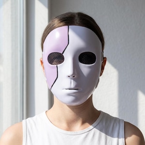 Könnte beinhalten: Eine glatte Vollmaske mit glänzend weißer Oberfläche und einem markanten lavendelfarbenen Bereich auf der linken Seite. Die Maske verfügt über dunkle, netzartige Augenöffnungen und eine dünne, schwarze vertikale Linie zur Trennung.
