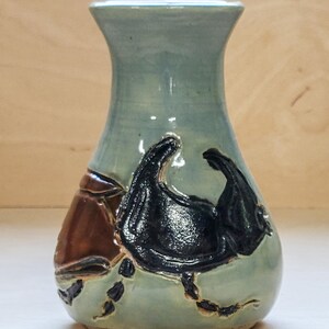 May include: A ceramic vase with a light blue-green glaze featuring a raised, dark brown and black relief sculpture of a beetle. The vase has a wide, rounded base that tapers upward to a flared rim, showcasing a smooth, glossy finish on the body.
