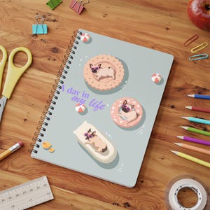 May include: A spiral-bound notebook with a light grey cover featuring three illustrations of pugs wearing pink sunglasses, each resting on a different pool float, with the text A day in my life printed in purple script across the centre.