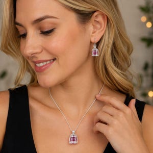 May include: A silver-toned jewelry set consisting of a pendant necklace and matching drop earrings. Each piece features a small, intricate cage-like design housing a vibrant, deep pink oval-shaped gemstone suspended from a delicate silver chain.