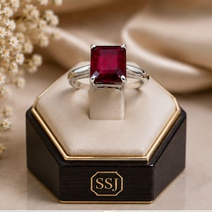 May include: A silver ring featuring a large, rectangular, deep red gemstone set in a four-prong setting. The ring is displayed on a beige, hexagonal stand with a dark brown base that features the gold-coloured letters SSJ in a rectangular border.