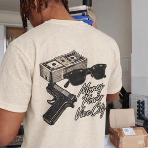 May include: A cream-colored speckled t-shirt featuring a black graphic on the back. The design includes a stack of cash, black sunglasses, a handgun, and the text "Money Power Vice City" written in a cursive script font.