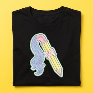 Könnte beinhalten: Ein schwarzes T-Shirt mit einer Stickerei eines gelben Bleistifts mit rosa Radiergummi und rosa Schleife. Ein langer, fließender Pferdeschwanz aus violettem und blauem Haar erstreckt sich vom oberen Ende des Stifts, umrandet von einem hellen Mintgrün.