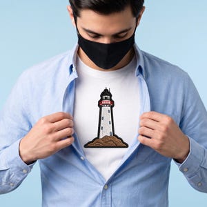 Könnte beinhalten: Ein weißes T-Shirt mit einem mittig platzierten, gestickten Aufnäher eines Leuchtturms. Der Leuchtturm ist weiß mit rotem Geländer und schwarzer Spitze und steht auf einem braunen Felsensockel. Das T-Shirt wird unter einem hellblauen Hemd getragen.