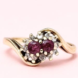 May include: A gold bypass ring featuring two round-cut deep red rubies at the centre, surrounded by a cluster of small, clear, round-cut diamonds. The band has a smooth, polished gold finish that curves around the central gemstone setting.