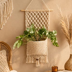 Puede incluir: Un tapiz de macramé color crema con una sección superior de patrón de diamantes y un bolsillo de tejido sólido en la parte inferior. La pieza cuelga de un palo de madera con un cordón e incluye una cuenta de madera decorativa en el fleco.