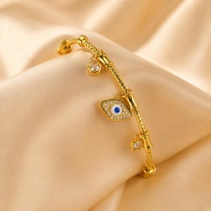 May include: A gold-toned bracelet featuring a textured chain with a central eye-shaped charm containing a blue and white pupil, surrounded by clear pavé stones, and accented with two additional small circular charms set with clear stones.