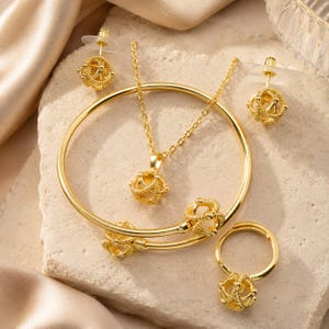Könnte beinhalten: Ein goldfarbenes Schmuckset bestehend aus einer Halskette, einem Armreif, einem Ring und einem Paar Ohrsteckern. Jedes Teil verfügt über einen kugelförmigen, käfigartigen Anhänger mit einer strukturierten, detaillierten Oberfläche.