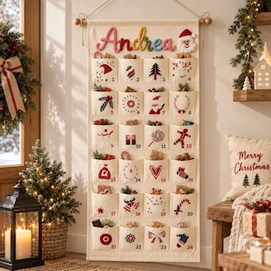 Pode incluir: Um calendário do advento de tecido cor de creme pendurado numa haste de madeira, com o nome Andrea em letras de lã coloridas no topo. Possui vinte e quatro bolsos numerados, cada um decorado com ícones de Natal vermelhos e pretos.