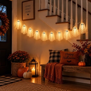 May include: A string of eleven illuminated ghost-shaped lights with black eyes, each featuring a textured, semi-transparent white finish. The string lights are draped horizontally, emitting a warm glow against a neutral wall.