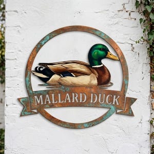 May include: A circular metal wall sign with a weathered copper and teal patina finish. The center features a detailed illustration of a mallard duck. Below the duck, a banner displays the text MALLARD DUCK in bold, capital letters.