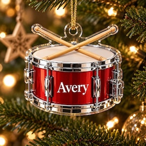Könnte beinhalten: Ein roter Snare-Drum-Weihnachtsschmuck mit einem weißen Trommelfell und zwei gekreuzten hölzernen Trommelstöcken oben. Die Seite der Trommel ist metallisch rot und trägt den Namen Avery in weißen Buchstaben. Mit goldener Kordel.